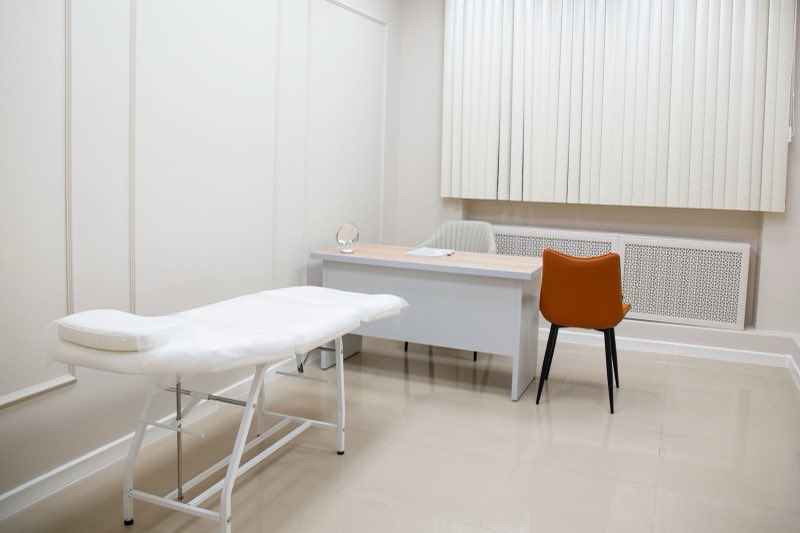 An empty consulting room with a treatment couch on the left, a plain desk and a single chair against the far wall.
