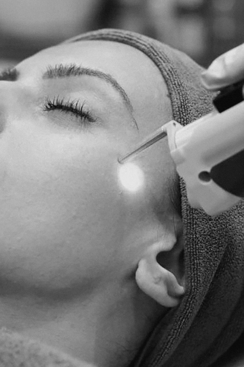 Black and white photograph of a handheld device held against a cheek, its tip lit, with a towel wrapped around the hair and the eyes closed.