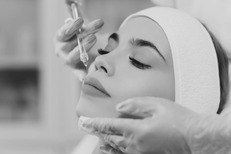 Black and white photograph of a gloved hand holding a fine needle above the upper lip, the other gloved hand steadying the chin.