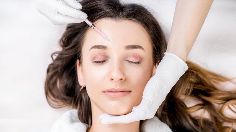 Photograph looking down at a person lying back with their hair fanned out, a gloved hand bringing a fine syringe towards the forehead and the other steadying the jaw.
