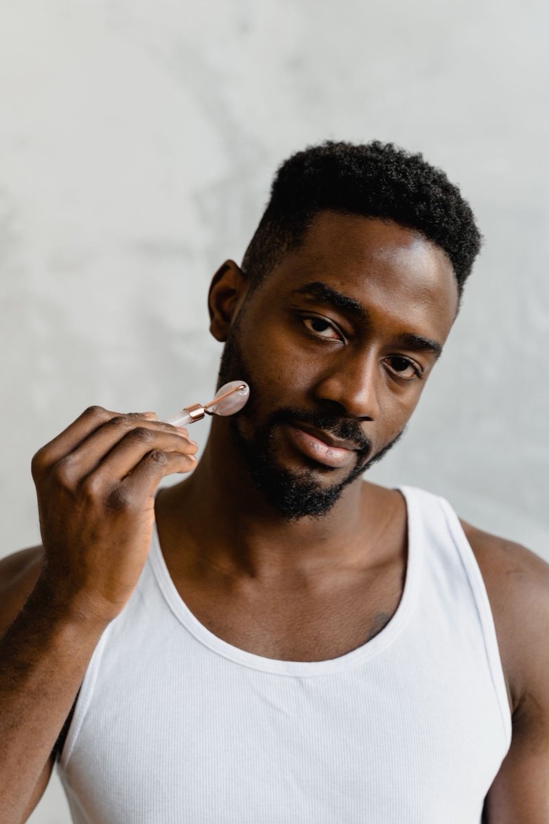 A man in a plain vest standing against a bare wall, holding a small facial roller to his cheek and looking at the camera.
