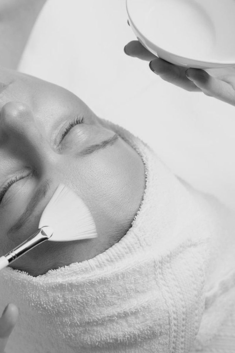 Black and white photograph of a treatment mask being spread across a cheek with a wide fan brush, a small white bowl held above the frame.