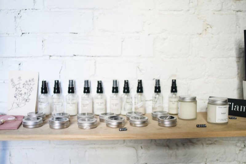 A row of plain pump bottles and small metal tins lined up on a wooden shelf against a painted white brick wall.