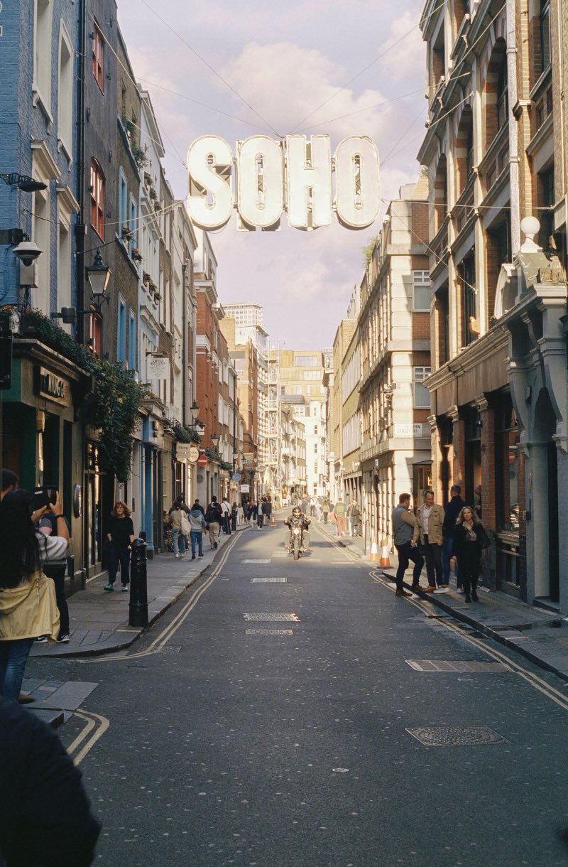 A narrow central London street of shopfronts and upper storey windows, with people walking in the road and a large SOHO sign strung overhead.